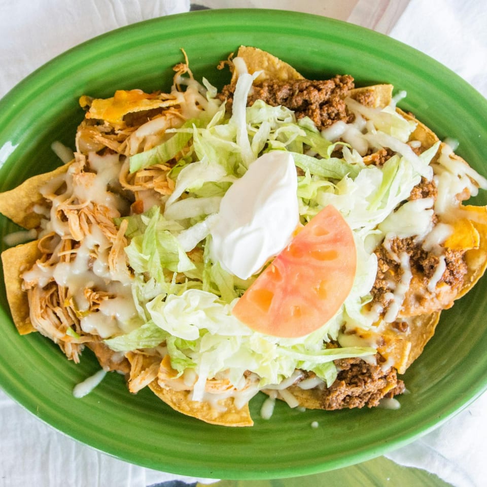 Palacio Maya's nachos with shredded chicken, ground beef, lettuce, sour cream, and tomato on a green plate.