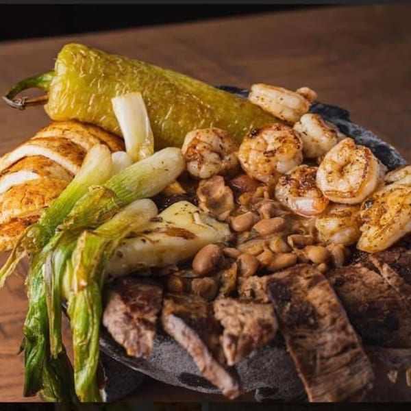 A molcajete filled with grilled shrimp, steak, beans, green onions, and a large green chili.