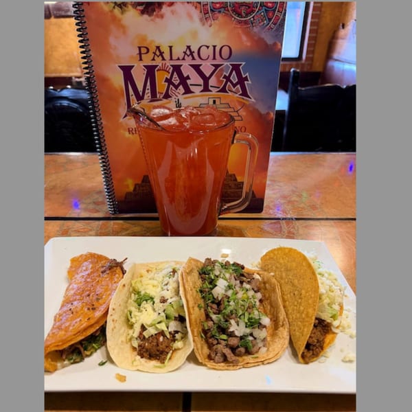 A plate of four diverse tacos and a red drink with ice, with a Palacio Maya menu in the background.